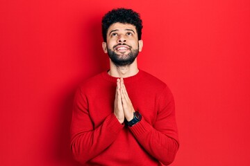 Young arab man with beard wearing casual red sweater begging and praying with hands together with hope expression on face very emotional and worried. begging.