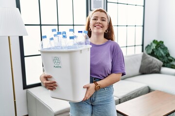 Young redhead woman holding recycling wastebasket with plastic bottles with a happy and cool smile on face. lucky person.