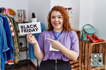 Young redhead woman holding black friday banner at retail shop smiling happy pointing with hand and finger