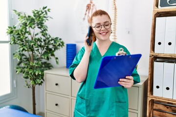 Young redhead woman wearing phsiologist uniform talking on the smartphone at clinic