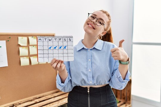 Young redhead woman holding travel calendar at the office smiling happy and positive, thumb up doing excellent and approval sign
