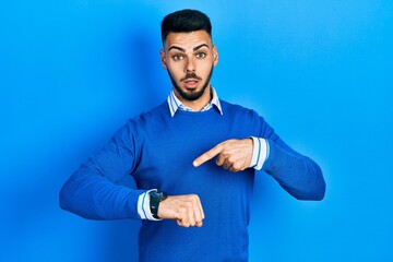 Young hispanic man with beard wearing casual blue sweater in hurry pointing to watch time,...
