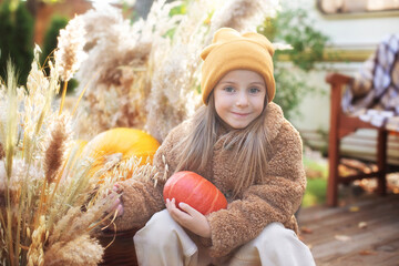 happy child with pumpkin outdoor in halloween in garden. Smiling girl sit on porch with a pumpkin in her hands. Trick-or-treat. Happy little girl playing on steps home. Child in cozy fall backyard. 