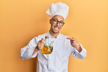 Bald man with beard wearing professional cook apron holding olive oil smiling happy pointing with hand and finger