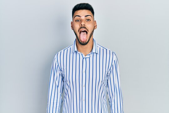 Young hispanic man with beard wearing casual striped shirt sticking tongue out happy with funny expression. emotion concept.