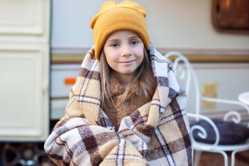 Smiling young girl with warm blanket standing on porch RV house in fall garden. Child covered with blanket on terrace house to warm up. Child girl in campsite backyard. Concept camping, adventure 
