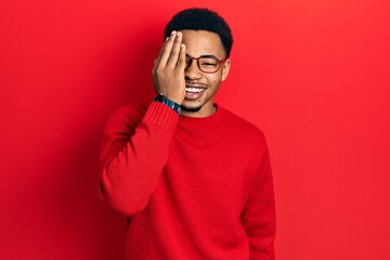 Young african american man wearing casual clothes and glasses covering one eye with hand, confident smile on face and surprise emotion.