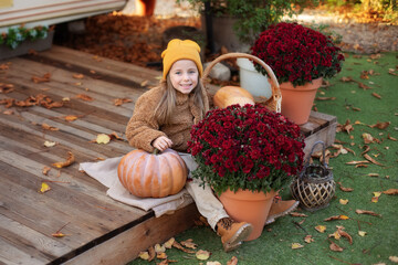Happy little girl sitting on porch of house with chrysanthemum potted and pumpkins. home fall...