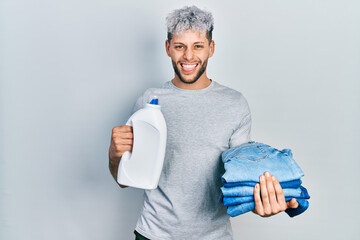 Young hispanic man with modern dyed hair holding jeans for laundry and detergent bottle sticking tongue out happy with funny expression.