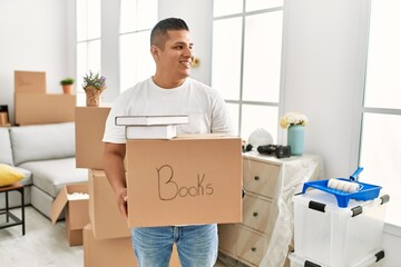 Young latin man smiling happy holding books cardboard box moving at new home.
