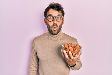 Handsome man with beard holding peanuts scared and amazed with open mouth for surprise, disbelief face