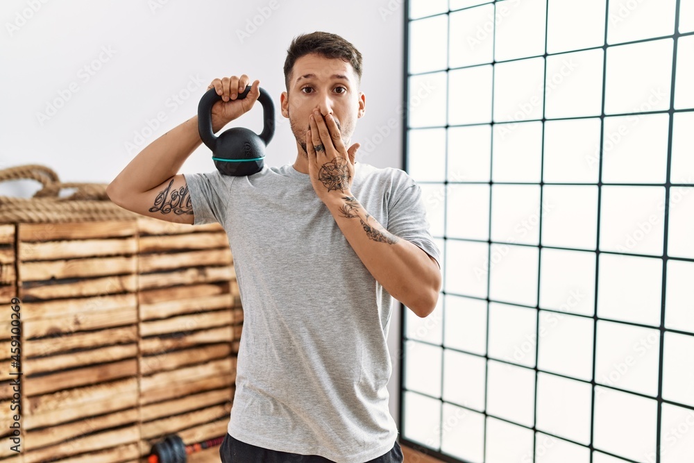 Wall mural Young handsome man wearing sportswear using dumbbells covering mouth with hand, shocked and afraid for mistake. surprised expression