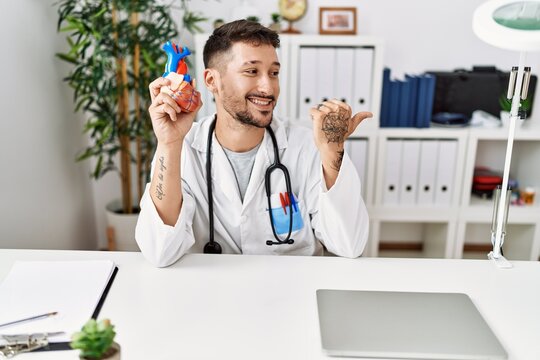 Young doctor holding heart at medical clinic pointing thumb up to the side smiling happy with open mouth