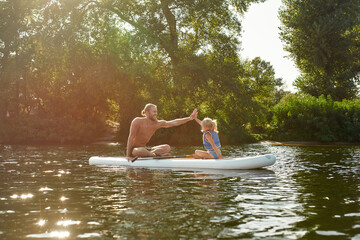 Father giving high five to his little daughter while relaxing, sup surfing on a river surrounded by the beautiful nature on a summer day