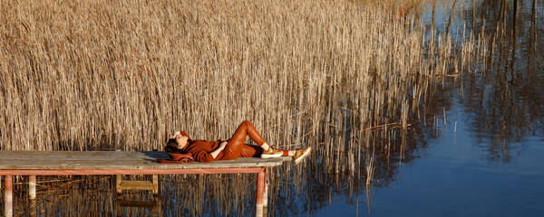 Portrait of a woman in autumn cozy orange clothes with backpack sitting on a wooden bridge near the lake with blue water and looking agains the sun. People, travel, nature concept.
