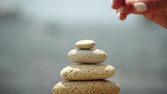 Woman bilds stones pyramid on the seashore on a sunny day on the blue sea background. Happy holidays. Pebble beach, calm sea, travel destination. Concept of happy vacation on the sea, meditation, spa