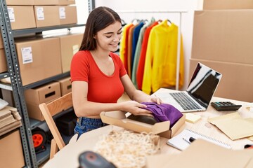 Young hispanic woman smiling confident packing clothes box at warehouse