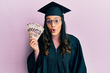 Young hispanic woman wearing graduation uniform holding mexican pesos banknotes scared and amazed...
