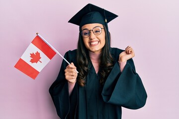 Young hispanic woman wearing graduation uniform holding canada flag screaming proud, celebrating victory and success very excited with raised arm