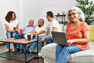 Fototapeta premium Group of middle age friends sitting on the sofa speaking. Woman smiling happy using laptop at home.