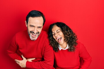 Middle age couple of hispanic woman and man hugging and standing together smiling and laughing hard out loud because funny crazy joke with hands on body.