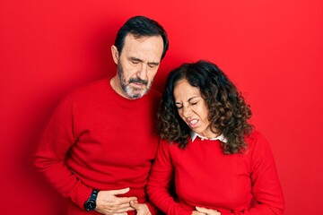 Middle age couple of hispanic woman and man hugging and standing together with hand on stomach because indigestion, painful illness feeling unwell. ache concept.