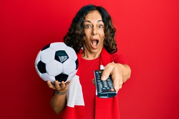 Middle age hispanic woman football hooligan holding ball using tv control celebrating crazy and...