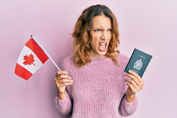 Young caucasian woman holding canada flag and passport angry and mad screaming frustrated and...
