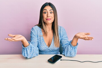 Young hispanic woman wearing casual clothes charging smartphone battery clueless and confused...