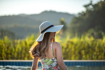 Back view of a young Asian woman with long hair wearing white straw hat relaxing in warm summer swimming pool on a sunny day. Tropical summer vacation and holliday concept