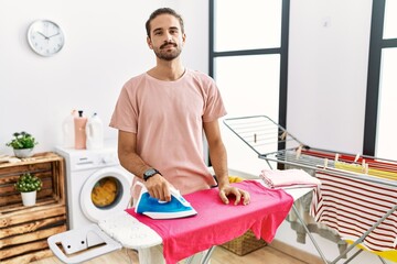 Young hispanic man ironing clothes at home relaxed with serious expression on face. simple and natural looking at the camera.