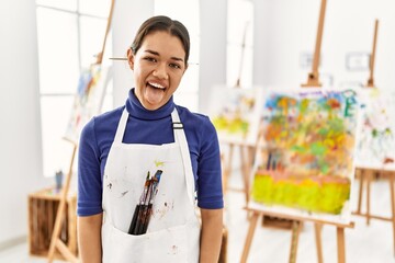Young brunette woman at art studio sticking tongue out happy with funny expression. emotion concept.