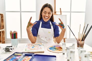 Young brunette woman at art studio with painted hands shouting with crazy expression doing rock...