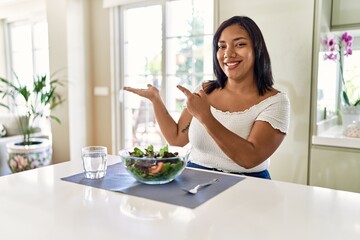 Obraz na płótnie Canvas Young hispanic woman eating healthy salad at home amazed and smiling to the camera while presenting with hand and pointing with finger.