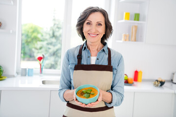 Photo of mature sweet lady hold soup wear apron jeans shirt at kitchen alone