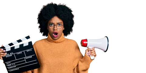 Young african american woman holding video film clapboard and megaphone afraid and shocked with...