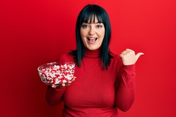 Young hispanic woman holding bowl of pills pointing thumb up to the side smiling happy with open mouth