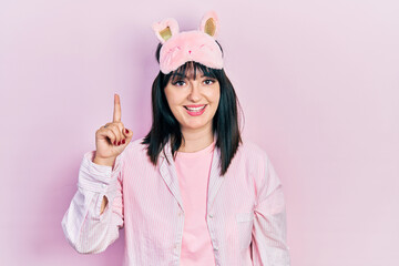 Young hispanic woman wearing sleep mask and pajama pointing finger up with successful idea. exited and happy. number one.