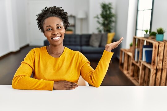 Young african american woman wearing casual clothes sitting on the table at home smiling cheerful presenting and pointing with palm of hand looking at the camera.