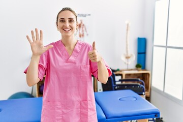 Young blonde woman working at pain recovery clinic showing and pointing up with fingers number six while smiling confident and happy.