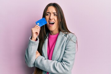 Young hispanic girl holding credit card angry and mad screaming frustrated and furious, shouting...