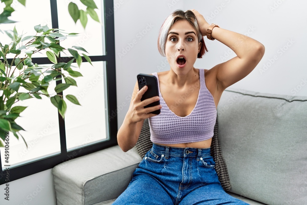 Sticker Young beautiful woman using smartphone typing message sitting on the sofa crazy and scared with hands on head, afraid and surprised of shock with open mouth