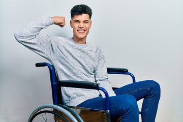Young hispanic man sitting on wheelchair strong person showing arm muscle, confident and proud of...