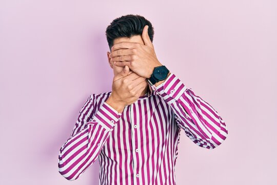 Young hispanic man wearing casual clothes covering eyes and mouth with hands, surprised and shocked. hiding emotion