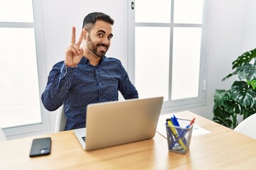 Young hispanic man with beard working at the office with laptop smiling looking to the camera showing fingers doing victory sign. number two.