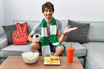 Young hispanic man football hooligan cheering game at home pointing aside with hands open palms showing copy space, presenting advertisement smiling excited happy