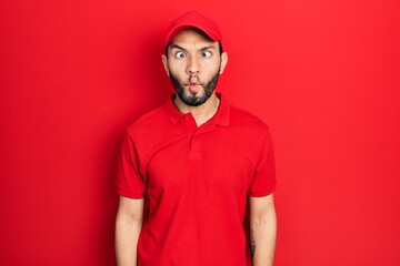 Hispanic man with beard wearing delivery uniform and cap making fish face with lips, crazy and comical gesture. funny expression.