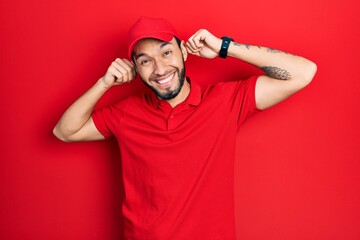 Fototapeta premium Hispanic man with beard wearing delivery uniform and cap smiling pulling ears with fingers, funny gesture. audition problem