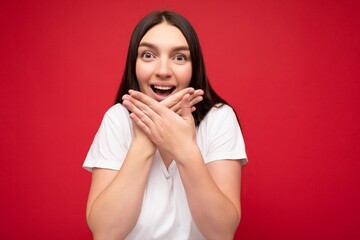 Photo of positive young beautiful brunette female person with sincere emotions wearing casual white t-shirt for mockup isolated over red background with copy space and covering mouth