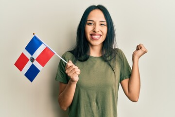 Young hispanic girl holding dominican republic flag screaming proud, celebrating victory and...